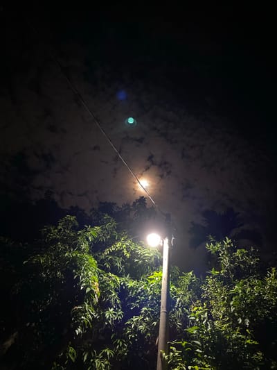 A night scene in Lambaro Kueh, Indonesia, features a street lamp illuminating lush green foliage. The sky above is dark, with scattered clouds, and a faint moon is visible through the clouds. A power line runs diagonally across the upper portion of the image. The street lamp is positioned on the right side, with its light directed downwards, casting a warm glow on the surrounding trees. The overall atmosphere is serene and tranquil, typical of a quiet evening in a residential area.