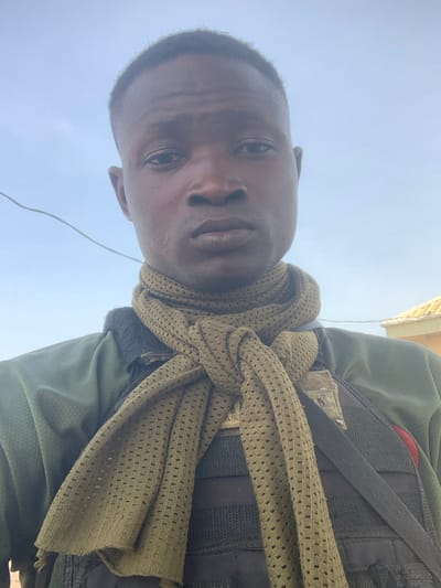 A selfie of a dark-skinned man with short hair, appearing to be outdoors under a blue sky. He's wearing military-style attire including a vest with straps and an olive green, mesh scarf wrapped around his neck. The camera angle is low, pointing up at his face. His expression is neutral.