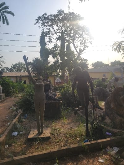 The scene depicts an outdoor sculpture garden in San Martín, Colombia. In the foreground, two human-figure sculptures are visible on dry, uneven ground. The left sculpture is a light-colored, nude female figure standing upright on a square base, with both arms raised vertically overhead. The right sculpture is a darker-toned figure, possibly male, depicted in a dynamic, forward-leaning pose with one leg lifted. The ground consists of sparse grass and dirt, contained by low concrete curbs, and is littered with various debris including plastic bags and bottles. In the mid-ground, additional concrete sculptures, including a reclining figure on a pedestal and an animalistic form, are partially visible among overgrown shrubs. The background features several trees, including a large deciduous tree with dense foliage and tall, slender trees, partially obscuring yellow-painted buildings with red roofs. Multiple utility lines traverse the upper portion of the frame against a clear sky, where a bright sun flare originates from the upper right, indicating strong direct sunlight.