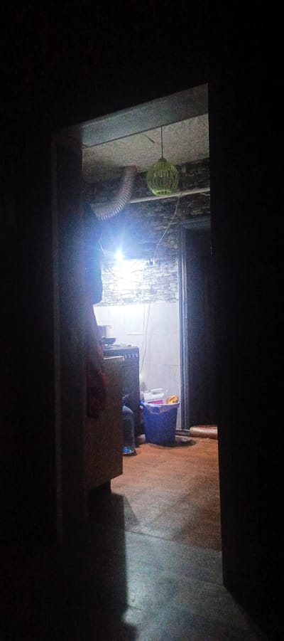 The image shows a dimly lit interior space, possibly a kitchen or utility room, viewed through an open doorway. A green, orb-shaped light fixture hangs from the ceiling in the upper right portion of the frame. In the background, a stove is visible, with a bright light source illuminating the area above it. To the right of the stove, a blue plastic bucket is present, containing various items, including a white container with a pink lid and what appears to be a yellow object. A person, whose lower body is visible, is seated in front of the stove, wearing blue trousers and a dark top. The floor is made of concrete and shows signs of wear. The walls have a tiled or brick-like pattern. The overall impression is one of a functional, but somewhat cluttered, domestic space. The location is identified as Khartsyzk, Ukraine.
