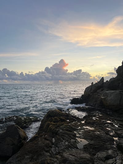 This image captures a serene sunset over the ocean at a rocky coastline, likely in Choeng Thale, Thailand. The sky is painted with soft hues of blue, pink, and orange, with wispy clouds scattered across the horizon. The sea is calm, with gentle waves lapping against the shore. In the foreground, dark, jagged rocks are covered in patches of seaweed and small tide pools, reflecting the colors of the sky. Two figures are visible on the rocks, appearing to be fishing. One is sitting down, while the other stands near a fishing rod. The scene evokes a sense of peace and tranquility as day transitions into night.