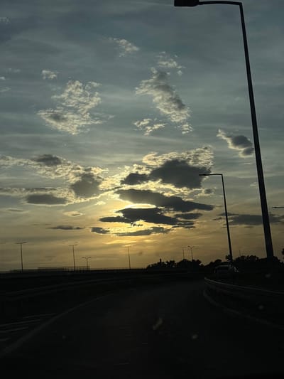 A landscape shot from a moving vehicle, likely a car, on a roadway in Obrenovac Urban Municipality, Serbia. The image is oriented sideways. The right side of the frame shows a dark asphalt road with a curved shoulder. The left side displays a sky filled with cumulus clouds, varying from white to dark gray, illuminated by the setting sun that casts golden rays. In the distance, evenly spaced streetlights stand along the road's edge. Overhead powerlines are visible against the sky. The shot appears to be taken during the sunset.