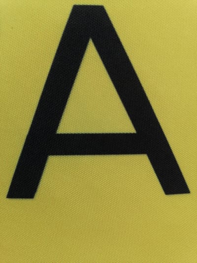 The image shows a close-up of a large, black letter "A" printed on a yellow textured fabric. The fabric appears to be made of a woven material, possibly canvas or a similar synthetic textile. The letter "A" is centrally located in the frame and dominates the image. Its edges are sharp and well-defined against the yellow background. The texture of the fabric is visible throughout the image, with small, repeating patterns of interwoven threads creating a grid-like effect. There are no discernible elements in the image that would suggest a location in the city of Durankulak, Bulgaria, or any other specific geographic setting. The image is devoid of any people, actions, or interactions.