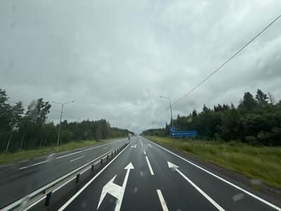 The image depicts a wet, overcast day on a multi-lane highway in Glukhaya Lokhta, Russia. The perspective is from inside a vehicle, looking forward. The asphalt road is marked with white lane lines and directional arrows indicating multiple lanes and turns. On the right side of the road, a blue sign with white text reads "ГЛУХАЯ ЛОХТА 0.4," indicating the location and distance to Glukhaya Lokhta. Dense green forest borders the highway on both sides, with tall trees and undergrowth. Streetlights are visible along the roadside. The sky is heavily clouded, suggesting rain or impending precipitation, as indicated by droplets on the visible windshield. There are no people or other vehicles immediately discernible in the foreground or mid-ground, though a distant vehicle is visible on the highway. The scene conveys a sense of travel on a relatively empty highway under subdued weather conditions.