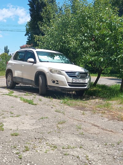 A white Volkswagen Tiguan SUV is parked on a cracked asphalt surface near a tree. The vehicle's license plate reads "P201HT 193 RUS". The scene is outdoors with a blue sky, white clouds, and lush green trees in the background. The ground is covered with patches of grass and small stones. The location is identified as Krymsk, Russia.
