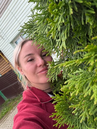 A woman with blonde hair and light skin is partially obscured by a dense, green conifer tree. She is looking at the camera with a slight smile, her face illuminated by natural light. She is wearing a red hooded sweatshirt and a black beaded necklace. In the background, a light-colored building with horizontal siding and a window is visible, along with wooden fencing and some green grass. The scene appears to be outdoors, possibly in a residential area of Chastyye, Russia, given the architectural style of the building and the presence of a conifer tree.