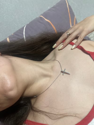 The image shows a close-up, high-angle view of a person lying on a pillow. The person has long, dark hair that partially covers their face and chest. They are wearing a red top and a silver necklace with a cross pendant. A ring is visible on their right hand, which is resting on their chest. The background consists of a gray wall and a pillow with a striped pattern. There is no indication of the location within the city of Postoyata, Ukraine.