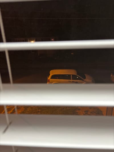 The image depicts a nighttime scene viewed through a window partially obscured by white vertical blinds. The primary subject is a white car, possibly a sedan or coupe, seen parked outside through the gaps in the blinds. Its rear section is most prominent, revealing a red taillight glowing faintly. Reflections from inside the car or a nearby light source are also visible on its surface.
The setting is dark, indicating it is night or late evening. The background is largely undifferentiated black, with a few faint light sources visible in the distance, possibly from other buildings or streetlights. To the right of the car, a textured surface, which appears to be pavement or a driveway, can be seen between the blind slats. The image itself is oriented vertically, suggesting it has been rotated 90 degrees clockwise from its original capture orientation. There are no people visible, nor is any discernible text present in the image. The scene is located in Franklin Township, United States of America.