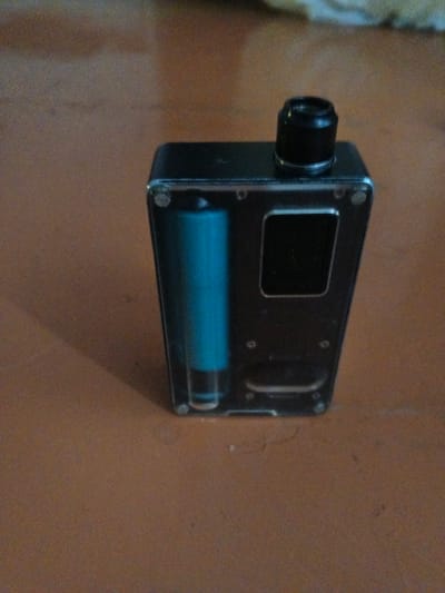 The image shows a close-up, high-angle shot of a black electronic cigarette device with a clear side panel, revealing a blue cylindrical battery inside. The device is positioned on a brown, textured surface that appears to be a table or floor. A black mouthpiece is visible on the top of the device. The background is slightly blurred, suggesting a shallow depth of field. There are no people or animals present in the image. The lighting appears to be from above, casting a subtle shadow to the left of the device. No specific details about the location in Artemovsky, Russia, can be discerned from the image itself.