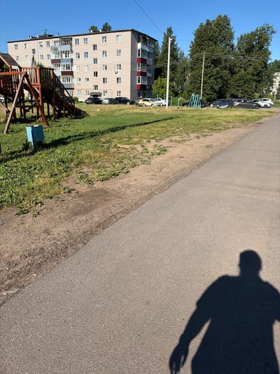 A shadow of a person, facing left, is cast on a paved asphalt path in the foreground of the image. The path runs diagonally from the bottom right to the top left. To the left of the path is a strip of dirt and grass. In the background, a multi-story apartment building with light-colored brickwork and balconies is visible. A children's playground structure, made of wood, is located to the left of the building. Several cars are parked in front of the building and along the side of the path. Tall green trees are situated behind the apartment building and playground. The sky is clear and blue. The scene appears to be a residential area in a city, likely on a sunny day.