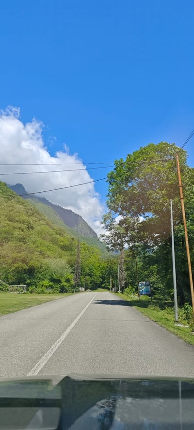 The image presents a first-person view from inside a vehicle, looking forward along a paved road winding through a scenic mountainous and forested landscape. The asphalt road features a clear white solid line down its center.

On both sides of the road, dense green trees and vegetation line the path. In the distance and background, steep, verdant mountains rise, their upper slopes partially obscured by large white clouds against a vibrant blue sky. Utility poles and power lines are visible, with a prominent tall wooden pole situated on the right side of the road. Another thinner, lighter-colored pole stands further along on the right. A blue sign with white text and graphics is partially visible on the right, nestled among the trees.

The scene is captured on a bright, sunny day, likely late morning or early afternoon, with scattered clouds. Reflections of the sky, clouds, and surrounding foliage are visible on the vehicle's dashboard and windshield at the bottom of the frame. The overall impression is one of a journey through a serene natural environment, typical of mountainous regions like Bzypta, Abkhazia. The text on the blue sign is not clearly legible.