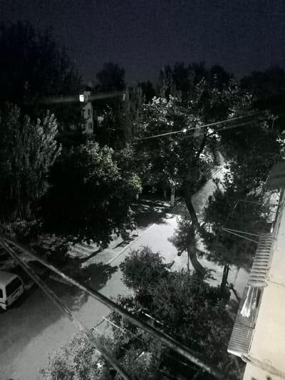 This is a nighttime, high-angle, black-and-white photograph taken from a building in Kokand, Uzbekistan. The scene depicts a residential street with trees lining both sides. A white van is parked on the left side of the street. A building with a lit window is visible in the background, partially obscured by trees. Power lines stretch across the image from the foreground towards the center. The street appears to be paved, and a park bench is visible on the right side of the street. The overall atmosphere is dimly lit and somewhat obscured by darkness and the presence of trees.
