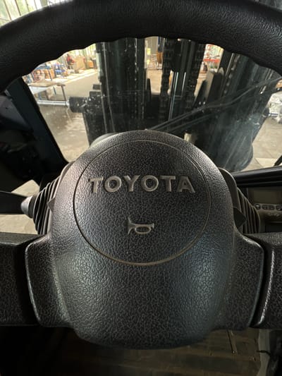 The image shows the interior of a Toyota forklift, focusing on the steering wheel which has the word "TOYOTA" embossed on it. A horn symbol is also visible on the steering wheel. The background shows the interior of a warehouse or industrial setting with light filtering in from windows. Forklift masts and other equipment are visible in the background. The location is the city of Lanžhot, Czechia.