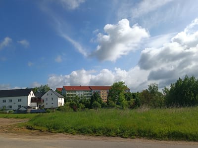 The image shows a landscape in Lunzenau, Germany, featuring a field of green grass in the foreground. A section of a road or pavement is visible in the bottom of the image. Behind the field, there are several houses. One complex has a dark-colored roof and white walls, while further back there is a large residential building with a red roof and yellow and light green walls. A cluster of trees surrounds these buildings. The sky is mostly blue with many white clouds.