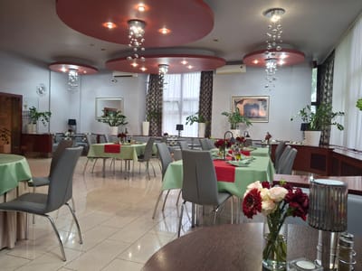 The image depicts the interior of a restaurant or dining hall, likely in Pila, Poland. The setting appears to be empty of people, suggesting it is either before opening hours, after closing, or during a quiet period.
The room features multiple dining tables, each covered with a light green tablecloth and a red runner. The tables are set with plates, napkins, and what appear to be condiments, indicating preparations for service. Several grey chairs with chrome legs surround the tables.
Dominating the ceiling are modern, circular red fixtures from which contemporary chandeliers with cascading glass orbs hang, casting a warm light. These are complemented by recessed ceiling lights. Large windows draped with sheer white curtains are visible, suggesting a bright daytime setting. Decorative elements include potted plants placed strategically around the room and framed artwork on the walls.
A close-up in the foreground reveals a floral arrangement in a glass vase, composed of red and cream roses and a dark red gerbera daisy. Next to it is a decorative cylindrical lamp with a reflective surface. The floor is tiled with a light-colored, large-format tile, which reflects the lighting and the overall ambiance of the space. The overall impression is one of understated elegance and readiness for patrons.