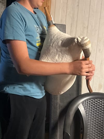 The image depicts a person in Sadovo, Bulgaria, holding a bagpipe. The person is wearing a blue t-shirt and black pants. They are holding the bagpipe with both hands. The bagpipe is made of white material and has a wooden mouthpiece. The background consists of a white wall with vertical slats and a black chair. The lighting in the image is dim.