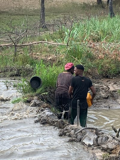 Two individuals stand along a muddy riverbank, with a black bucket positioned near the water's edge. The person in the foreground wears a pink head covering and a brown shirt. The other person is dressed in a black shirt and holds a yellow container. The river flows rapidly, causing splashes against the muddy bank, with vegetation growing on the opposite side. A thin tree with sparse branches is visible on the left of the frame.