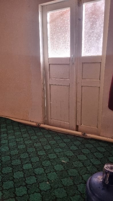 A grimy white-framed window with two panes sits above a peach-colored wall. Natural light filters through the obscured glass. To the right, a green and black patterned carpet extends, partially covered by a blue water jug. A thin, light-colored rod runs along the edge of the carpet, bordering the wall. The scene appears indoors and is slightly angled.