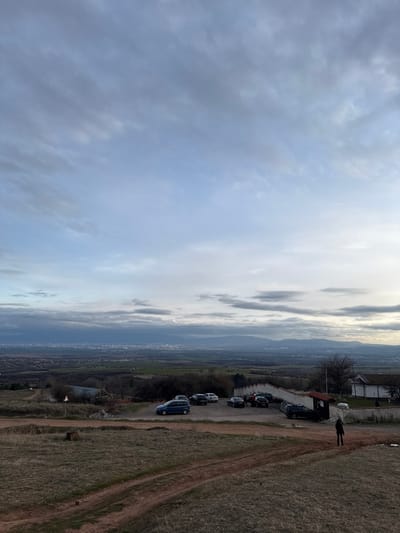 The image displays an elevated panoramic view from the area of Balsha, Bulgaria, looking out over a valley and a distant urban area. The sky, occupying the upper two-thirds of the frame, is largely overcast with layered grey and pale blue clouds.
The immediate foreground consists of dry, sparsely vegetated, yellowish-brown terrain marked by multiple reddish-brown dirt paths. In the midground, an unpaved parking area holds approximately eight vehicles, including a blue minivan and various dark-colored cars. To the right of the parking area, a section of a white-walled building with a dark roof is visible, enclosed by a low white wall. Further to the left and behind the parked vehicles, a smaller, dark structure is present among bare trees. One individual, dressed in dark clothing, is walking on a dirt path in the lower-right midground, moving away from the parked cars.
The background reveals a sprawling cityscape situated in a valley, partially obscured by atmospheric haze. Beyond the city, a range of mountains stretches across the horizon beneath the cloudy sky. The overall vegetation suggests a non-foliated season.