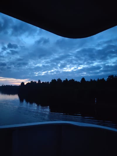 The image depicts a serene twilight scene over a body of water, likely a river or lake, with a silhouetted treeline bordering the far bank. The sky is a deep blue, filled with dramatic clouds that are tinged with the last remnants of sunset. The water reflects the moody sky and the dark shapes of the trees, with gentle ripples disturbing the surface. In the foreground, the edge of a dark structure, possibly the interior of a boat or a similar vessel, frames the view. The overall impression is one of tranquility and the approaching end of the day. The location is specified as Bessonovo, Russia, suggesting a natural landscape within or near the town. There are no people or specific actions visible in the image, only the quiet beauty of the natural environment at dusk.