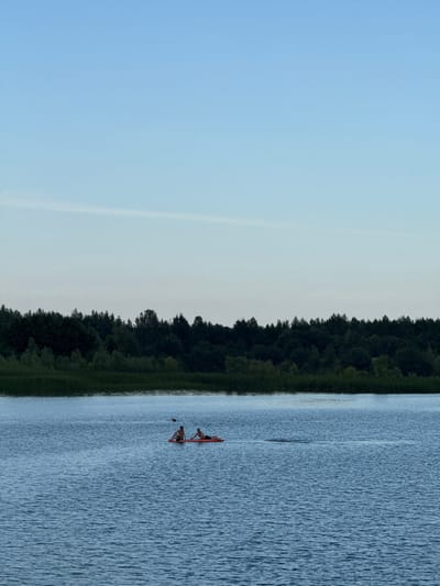The image depicts a serene natural scene at what appears to be dusk or early morning in Zaosinovo, Russia. The sky is a soft, gradient blue, transitioning to a lighter hue near the horizon, with a faint, wispy cloud formation visible. A dense line of dark green trees forms a natural border along the left side of the image, meeting a calm, blue body of water that occupies the right side.
On the water, two individuals are engaged in an activity on a small, red boat, possibly a kayak or canoe. One person is seated in the boat, while the other appears to be either partially in the water or interacting with the boat. The water's surface is rippled, reflecting the soft light. The overall atmosphere is tranquil and peaceful, suggesting a quiet moment spent outdoors. There are no visible text elements in the image.