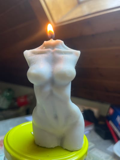 A white, torso-shaped candle with a lit wick is centered in the foreground of the image. The candle is resting on a bright yellow lid, which appears to be part of a container. The background is blurred, showing wooden ceiling beams and a hint of a window or skylight, suggesting an indoor setting possibly in an attic or room with sloped ceilings. There are some objects in the background that are out of focus, including what appears to be a stack of books or boxes and a red object. The lighting suggests it may be daytime, with light streaming in from above. This scene is located in Schwäbisch Hall, Germany.