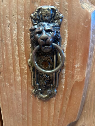 The image displays a close-up, vertical view of an antique-style metallic fixture mounted on a light-colored wooden surface, observed in Monterey, United States of America. The fixture, likely a decorative door pull or knocker, is crafted from tarnished dark bronze or brass. It features a prominent central lion's head, rendered with detailed facial features and mane, holding a movable, smooth circular metal ring in its open mouth. Above the lion's head, a smaller, lighter-toned brass or gold ornament depicts an ornate relief, possibly a human or cherub face framed by intricate elements. Below the lion, an elongated, ornamental backplate with scrolled patterns secures the fixture to the wood. A small, circular aperture is present in the backplate, positioned directly beneath the lion's mouth and within the metal ring. The backplate is secured by at least one visible flat-head screw at its lower end. The wooden background exhibits prominent vertical grain patterns and a natural finish, with minor surface texture inconsistencies and light wear. No individuals or active events are present within the scene.