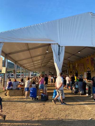 The image depicts an outdoor event under a large white tent structure. The ground is covered in dry earth and straw. In the foreground, a man in shorts and a white t-shirt walks away from the camera, his shadow cast on the ground. To his left, a blue shopping cart with the word "THERMO" is visible. Behind him, several people are seated on hay bales. Further into the tent, a long row of tables with people seated and standing is visible. A decorative yellow wall with shield motifs and writing, possibly a banner, is present in the background. The sky is clear and blue. Based on contextual information about the location, this scene appears to be at a festival or market in Llanera, Spain.