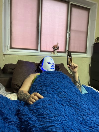 A person wearing a white LED facial mask with blue lights is lying on a couch, covered by a fluffy blue blanket. They are holding a smartphone in their right hand, which is illuminated by the phone's screen. The person has tattoos on their arms. Behind them is a window with pink blinds, and a small decorative arrangement of flowers in a vase sits on the windowsill. To the right of the person, a dark dresser is partially visible. The environment appears to be an indoor living space, likely a home in Murmansk, Russia, given the context.
