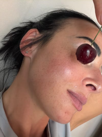 This media file is a close-up, overhead shot of a person's face, specifically the cheek, nose, and lips, with a cherry held near the eye. The person has dark hair and appears to be lying down, with their head resting on a light-colored surface. The lighting suggests an indoor setting, possibly a spa or a treatment room, with soft, diffused light.

The main subject is the person's face, which is captured in detail, highlighting the texture of the skin, including freckles and a slight sheen of moisture. A single cherry, with its stem, is positioned just below the person's right eye, held by a finger that is partially visible at the top of the frame. The cherry appears plump and dark red, with visible water droplets on its surface. The person's lips are parted slightly, revealing a pinkish tone. There is also a small stud earring visible on the person's earlobe.

No text is visible in the image. The scene is intimate and focused on the tactile and visual elements of the face and the fruit. The overall mood is calm and perhaps a bit surreal due to the unusual placement of the cherry. There are no clear indicators of time of day, weather, or specific location cues beyond the context provided. The activity taking place is unclear, but the focus on the face and the cherry might suggest a beauty treatment, a food-related sensory experience, or an artistic composition.