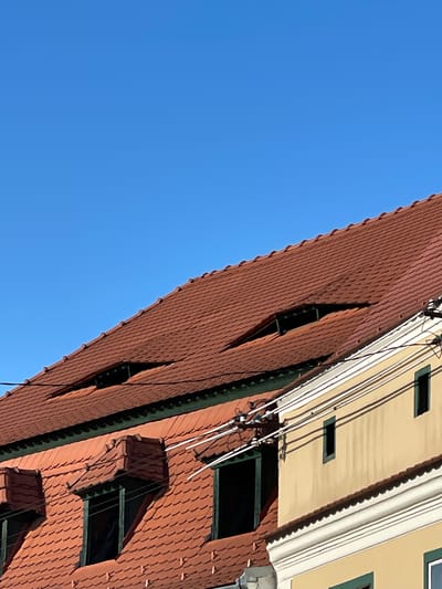 The image depicts the roof and upper portion of a building against a clear, blue sky. The building features a red tiled roof with several uniquely shaped dormer windows, some of which have dark green frames. A section of the building is painted beige with white architectural trim along the roofline. Electrical wires run along the building’s facade. This architectural style is characteristic of buildings in Sibiu, Romania.