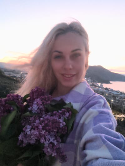A woman with blonde hair, fair skin, and light eyes is holding a bouquet of purple flowers. She's wearing a white and light purple plaid garment. The background shows a city on a coastline at dusk. The city is in Орджоникидзевский поселковый совет, Ukraine.