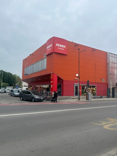 The image shows the exterior of a Penny Market supermarket, specifically the "Penny Autogara" location in Giurgiu, Romania. The building is primarily orange with red signage. A gray car is parked in front of the entrance, and several other vehicles are parked further down the street. Pedestrians are near the entrance, interacting with shopping carts. The sky is overcast. A street with a speed limit marked as "30" is adjacent to the supermarket. Trees are visible in the background.