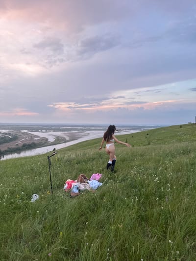In Britvino, Russia, a woman in white lingerie and black socks stands on a grassy hillside overlooking a river valley at dusk. A pile of clothing, a tripod, and a pink bag are on the grass nearby. The sky is filled with soft, pastel-colored clouds.