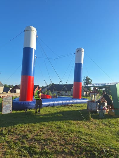 This image captures an outdoor event in Цімкавіцкі сельскі Савет, Belarus, on a clear day with a bright blue sky and a sliver of a moon visible. In the foreground, a large inflatable attraction, resembling two tall columns decorated with the colors of the Russian flag (red, white, and blue), is set up on a blue inflatable base. A man wearing a high-visibility yellow vest and camouflage pants stands near the inflatable structure, observing the scene. To the right of the attraction, a temporary canopy tent is erected, beneath which a woman in a black t-shirt and shorts is attending to a food stall. Several people are visible around the food stall, some appearing to be customers. The ground is covered in green grass, with some patches of exposed soil. In the background, several houses with brick and wooden exteriors are visible, suggesting a rural or semi-rural setting. The overall atmosphere appears to be a lively outdoor gathering or festival.