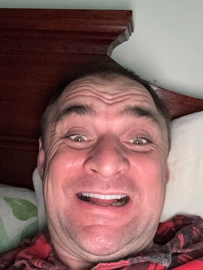 The image shows a close-up, selfie-style shot of a middle-aged man's face from the eyebrows down, with his mouth open in a wide smile, revealing his teeth. He is lying down, as indicated by the presence of a pillow and bedding. A wooden headboard is visible in the upper portion of the frame, and a wall is in the background. The man is wearing a red and black plaid shirt. The image appears to be taken indoors, likely in a bedroom. The facial features are prominent, with visible wrinkles around the eyes and across the forehead. The lighting suggests an indoor setting, possibly with artificial light. No specific landmarks or identifying features of Lesnoi Gorodok, Russia, are visible in the image.