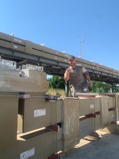 In Pankratove, Ukraine, under a clear blue sky, a man wearing a grey t-shirt, grey shorts, and a blue baseball cap stands in front of large, beige concrete blocks. He is making a "rock on" hand gesture and sticking out his tongue, appearing to be posing for the camera. The concrete blocks are arranged in a row, supported by metal beams and red brackets. Some of the blocks have warning labels with a lightning bolt symbol. In the background, more of the beige concrete elements are visible, stacked on an elevated metal structure. Trees and a fence line the edge of the scene. The overall environment suggests an outdoor industrial or construction site.
