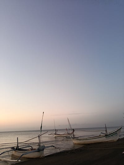 The image depicts a serene beach scene at sunrise or sunset in Jangkar, Indonesia. Several traditional Indonesian fishing boats, known as "prahu," are moored on the dark sandy shore and in the shallow water. The sky is a gradient of soft blues and pale oranges, indicating the time of day. The calm sea gently laps at the shore. In the foreground, a white prahu with outriggers is prominently displayed, showing some wear and tear. Another similar boat is partially visible to its left. Further in the distance, more boats are scattered along the coastline, some with masts and rigging. One of the boats in the mid-ground has the name "ALANDUTRA" written on its hull. The overall atmosphere is peaceful and tranquil, characteristic of a coastal fishing village.