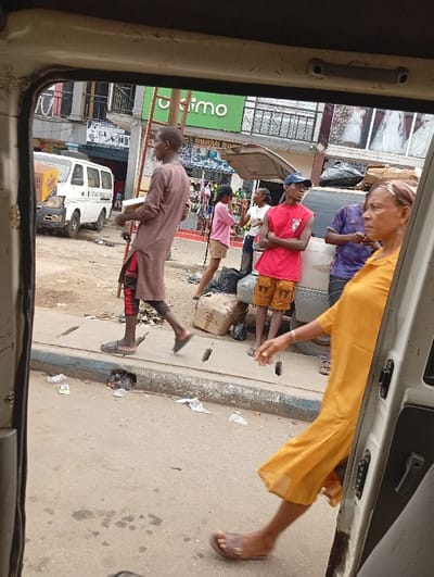 View from inside a vehicle showing a bustling street scene. Pedestrians stroll along the sidewalk next to a curb with litter. Several people are in view, including a man in a brown tunic, a woman in a pink shirt, a man in a red shirt and blue cap, and a woman in a yellow dress. A white van and other vehicles are parked nearby. A store with a green sign is in the background, alongside a building featuring a wedding dress display.
