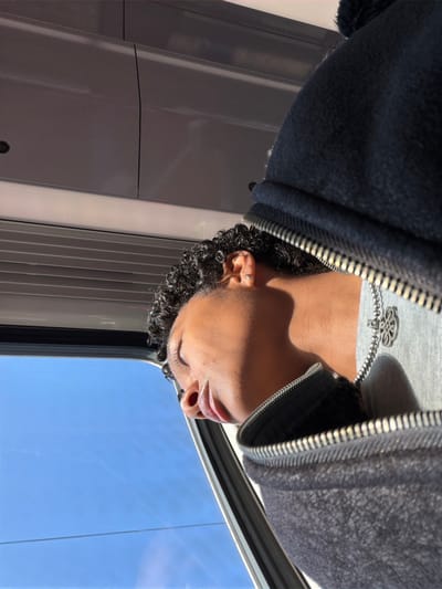 This image depicts a person inside a public transport vehicle in Chelles, France. The photograph is oriented 90 degrees clockwise from an upright perspective.
A single individual, with dark skin and dark, curly hair, is visible from the upper torso, with closed eyes, indicating a state of rest or sleep. They wear a dark, possibly navy or black, zippered hooded jacket, featuring a small dark pom-pom on the hood. A grey garment is visible underneath, complemented by a silver-colored chain necklace with a multi-lobed or floral pendant. A small silver-colored hoop earring is present in the helix of their right ear.
The person is positioned adjacent to a window. The vehicle's interior features grey overhead paneling and horizontal slatted structures below the window. Direct sunlight enters through the window, strongly illuminating the right side of the individual's face and jacket, casting pronounced shadows. The exterior view through the window shows an unobstructed clear blue sky. No other persons, objects, or specific contextual details of the exterior environment are visible.
