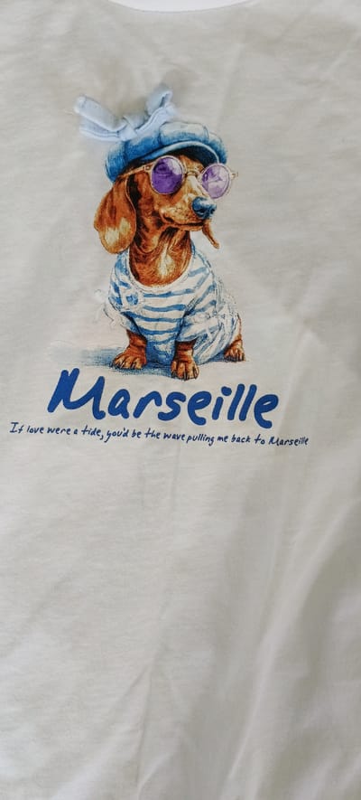 A white t-shirt features a graphic print of a dachshund wearing a blue cap and sunglasses, a striped shirt, and overalls. The word "Marseille" is printed in large blue cursive letters below the dog. Underneath, a smaller blue cursive phrase reads, "If love were a tide, you'd be the wave pulling me back to Marseille." The image shows a portion of the t-shirt, focusing on the printed design. The background is the white fabric of the t-shirt, with some wrinkles and folds visible. There are no elements in the image that indicate a specific location within Падлабенскі сельскі Савет, Belarus.