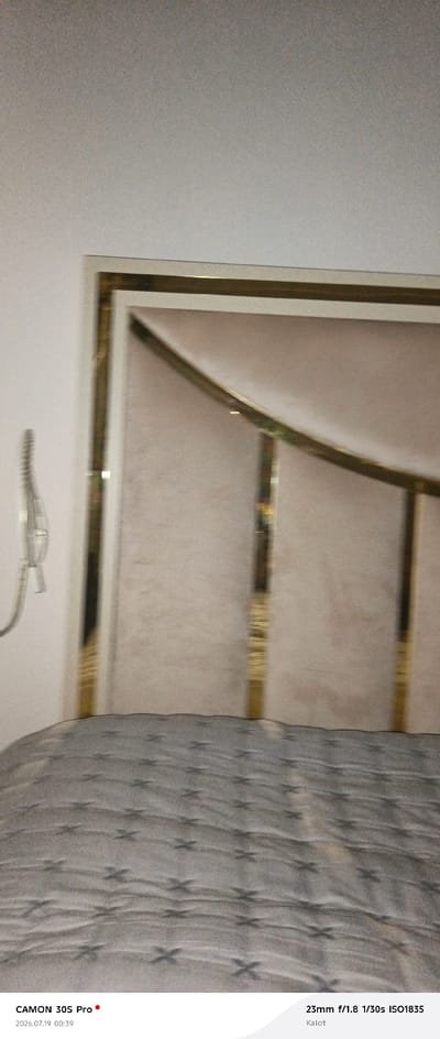 The image depicts a close-up view of a bed with a patterned duvet in the foreground. The duvet is a dark grey with a repeating pattern of stylized 'x' shapes in a lighter shade. The headboard of the bed is visible behind the duvet. It appears to be upholstered in a light-colored fabric, possibly beige or cream, and is framed by a decorative trim with a gold or brass-like finish. The trim has a geometric design, with straight and curved sections. To the left of the headboard, a wall-mounted item with a ribbed texture is partially visible. The lighting in the scene is soft, casting shadows that accentuate the texture of the duvet. The bottom edge of the image displays text indicating camera information: "CAMON 30S Pro" with a red dot, "2026.07.19 00:39" and "23mm f/1.8 1/30s ISO1835 Kalot". The location is identified as Kalot, Tajikistan.