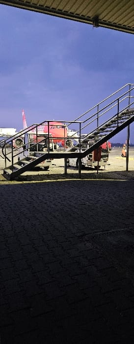 This image depicts an airport ramp area at Katowice Airport in Pyrzowice, Poland, during what appears to be twilight or early morning. The scene is viewed from beneath a light-colored, corrugated metal overhang, partially obscuring the top of the frame.
In the foreground, a dark, textured pavement surface, possibly interlocking pavers, is visible, partially illuminated by artificial light, casting subtle shadows.
The midground features a black metal staircase with multiple steps and handrails, angled upwards and to the right, extending out of the frame. Beyond the staircase, on a lighter tarmac surface, a red and white commercial aircraft is parked. The aircraft's fuselage is predominantly red with white sections, and a red tail fin with white markings is visible on the far left.
An adjustable passenger boarding stair unit, primarily white with silver steps and black railings, is positioned against the aircraft, aligned with a passenger door. Partially visible behind this unit is another piece of white ground support equipment. Further to the right, an orange ground vehicle or piece of equipment is discernible.
The background consists of a clear, dark blue sky, indicating ambient low-light conditions. No individuals are clearly visible within the frame. The aircraft appears stationary, with boarding stairs in place, suggesting preparation for or completion of passenger movement.