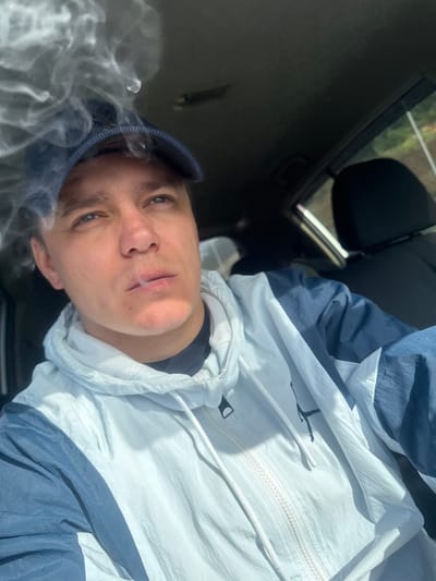 In Shkotovo, Russia, a man wearing a blue baseball cap and a blue and white windbreaker is captured in a car, looking upwards and to the right, with smoke billowing around his head and visible as it exits his mouth. The scene is lit by natural light, casting shadows and highlighting the texture of the windbreaker. The interior of the car is visible, with a dark headrest and window in the background, suggesting an outdoor setting.