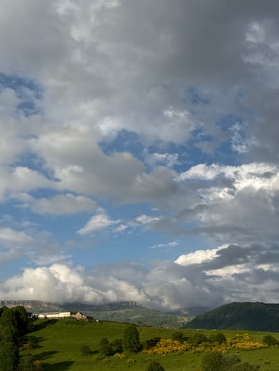 The image depicts a rural landscape with rolling green hills, a cluster of buildings on a hilltop, and a dramatic sky filled with clouds. In the foreground, the hills are covered in lush green grass with scattered trees and patches of yellow flowering bushes. A building complex, appearing to be a farm or a small settlement, is situated on the crest of the hill, with several structures visible, including what looks like a long, white-colored building and a smaller, darker-roofed building. In the background, distant mountains with flat, layered tops are visible, partly obscured by clouds. The sky is a dynamic mix of blue patches and various types of clouds, ranging from bright white cumulus to darker, more ominous formations, suggesting a changeable weather pattern. There are no people or animals clearly visible in the image. The scene is set in a natural, outdoor environment, likely in a mountainous or hilly region. The image is taken from a distance, capturing a wide view of the landscape.