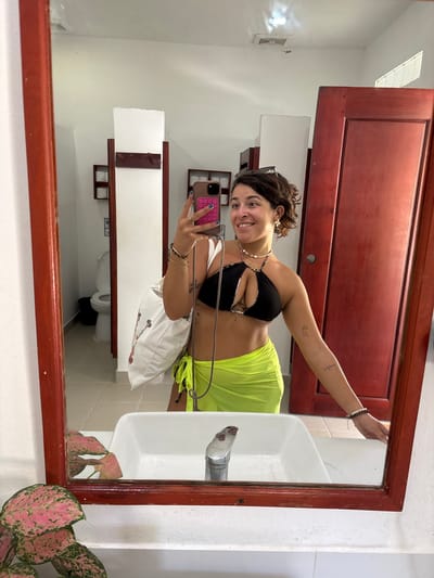 A young woman, facing a mirror, takes a selfie in what appears to be a public restroom. She is wearing a black bikini top and a neon green sarong. She has a white tote bag slung over her shoulder. The restroom has white tiled floors and walls, with wooden doors. A toilet and sink are visible in the reflection. A small lizard is perched on the ceiling in the reflection. The mirror is framed with reddish-brown wood. The overall impression is of a casual, travel-related setting.