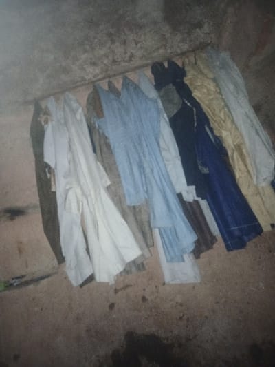 A collection of folded shirts lies on a textured, likely concrete floor. The shirts are various colors, including blue, brown, white, and khaki, suggesting a mix of formal and casual wear. The lighting is dim, with the shirts casting shadows, giving the scene a somewhat cluttered or disorganized impression. Some staining is visible on the floor.