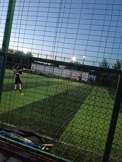 The image depicts an outdoor artificial turf soccer field at dusk. The sky is a gradient of pale blue to darker blue, indicating twilight. A chain-link fence with a green metal frame runs across the foreground, obscuring some of the view.
Several young people are on the field, appearing to be engaged in a soccer practice or game. One boy in a black shirt and shorts is prominently featured in the left-center of the image, facing away from the camera, with a bright yellow soccer cone near his feet. Further back on the field, other figures can be seen in athletic wear, some in motion.
In the background, there's a white building with a fence and trees. Lights are visible on poles, illuminating the field as the natural light fades. The overall atmosphere suggests a casual sporting event or training session taking place in the early evening. The weather appears clear. There is no visible text in the image.