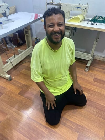 The image captures a man kneeling on a wooden floor in what appears to be a sewing workshop in Legenda, Egypt. He is wearing a bright yellow t-shirt and black pants. The man is smiling and looking directly at the camera. In the background, there are several sewing machines, work tables, and some fabric. One table has a JUKI brand sewing machine and a pair of glasses. Another table has a white surface with red and white striped tape that reads "ALL FOR FASTER SEWING". The overall environment suggests a functional workspace dedicated to garment production or alteration.