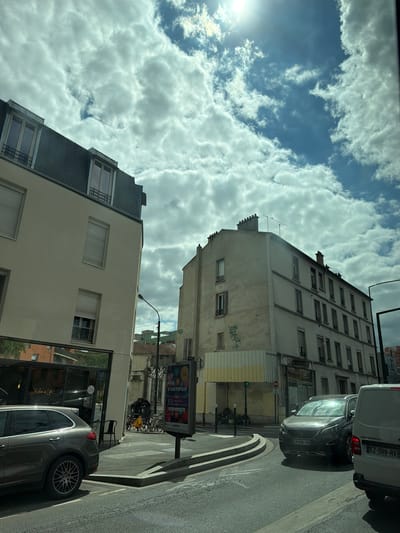 The image captures a sunlit street scene in Alfortville, France, under a partly cloudy sky. In the foreground, a dark grey SUV is stopped at a traffic light, with a white van to its right. The street features a curved road with white lane markings. To the left, a building with a light-colored facade and a dark sloped roof with dormer windows stands prominently. This building appears to house commercial spaces on the ground floor, with reflections of trees and buildings visible in its windows. A sign in the window reads "Chicken is life". A black chair sits on the sidewalk in front of the establishment. Further down the street, another building with a similar architectural style, featuring multiple windows, is visible. A streetlamp with a black pole stands near the sidewalk. A poster on a pole advertises "FEU D'ARTIFICE" (fireworks). Bicycles are parked near the entrance of the building to the left. The overall atmosphere suggests a typical urban environment during daylight hours.